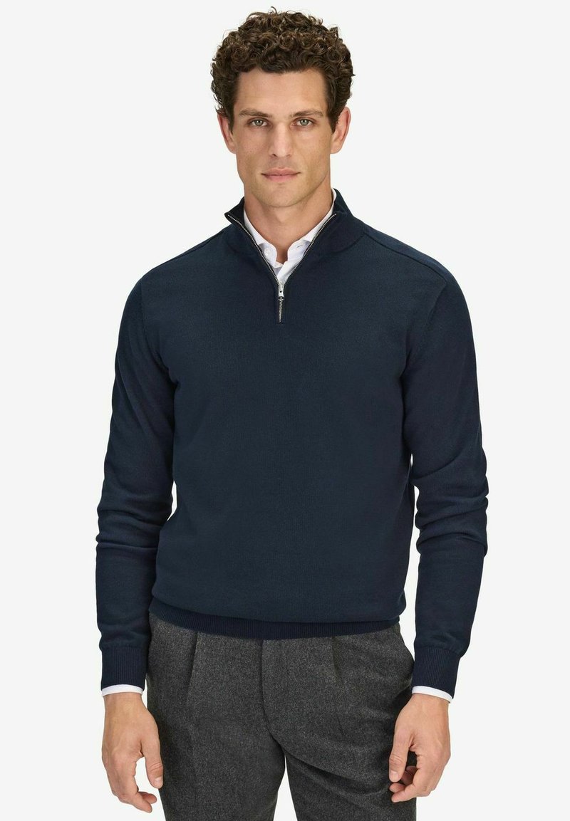 Tummansininen, vetskareilla varustettu puuvillaneulepullovaihtoehto. Varustettu kauluksella sekä resorilla varustetuilla hihoilla ja helmalla. Rikkaita valkoisen paidan päällä.