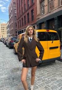 Blazer marrone oversize con maniche larghe, indossato sopra una camicia bianca e una cravatta blu scuro. Sullo sfondo ci sono un taxi giallo e edifici di mattoni.