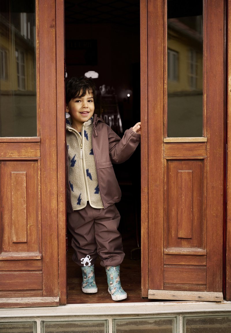 Bambino in piedi in una porta, indossa un giaccone impermeabile marrone, una felpa beige con design blu, pantaloni marroni coordinati e stivali da pioggia floreali.
