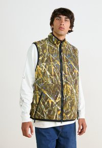 Fleece zip-up vest featuring a brown and yellow pattern resembling foliage. Contrast black zipper and trim. Worn over a white long-sleeve shirt.