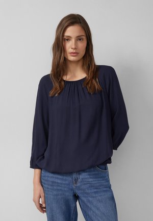 Vrouw met lang bruin haar, gekleed in een marineblauwe blouse met lange mouwen en een blauwe spijkerbroek, staand tegen een effen lichtgrijze achtergrond.