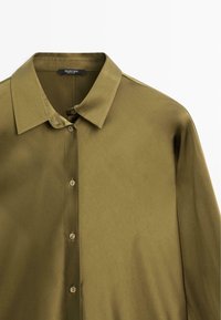 Camicia verde oliva abbottonata realizzata in tessuto morbido. Presenta un colletto a punta e cinque bottoni coordinati lungo la parte anteriore.