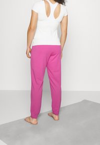 Kobieta stoi boso na szarej macie, ubrana w jasnoróżowe spodnie i białą krótką bluzkę z wycięciami na plecach.