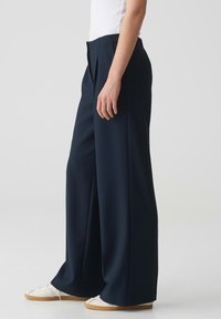 Brede marineblauwe broek van een gladde stof, met plooi(c)(en) bij de taille, gecombineerd met witte sneakers en een aansluitend wit topje.