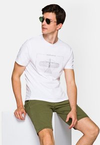 Biała bawełniana koszulka z nadrukowanym wzorem i tekstem, zestawiona z oliwkowymi szortami do kolan, noszona z okrągłymi czarnymi okularami przeciwsłonecznymi.