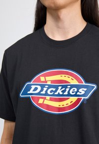 Svart bomullst-skjorte med et sirkulært logo med røde og gule detaljer og ordet "Dickies" i hvitt på et blått banner.