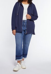 Marinblå huvtröja med dragkedja och dragskor, med en avslappnad passform. Matchas med blekta blå jeans och vita sneakers, vilket framhäver en avslappnad stil.