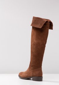 Brązowe zamszowe kozaki za kolano z zakładanym mankietem, okrągłym noskiem i niskim obcasem typu blok. Tekstura jest miękka o matowym wykończeniu.