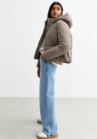 Pufferjack in gedempt taupe met een capuchon, voorzien van een gewatteerd ontwerp. Gecombineerd met lichtblauwe rechtgesneden jeans en beige instap-schoenen.