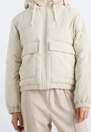 Beige corduroy pufferjack met voorzakken en elastische manchetten, gedragen over lichtbeige broek met fijne strepen.
