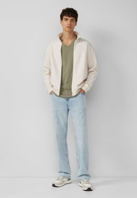 Jasny, zapinany na zamek kurtka na zielonym t-shircie z dekoltem w serek, zestawiona z jasnoniebieskimi dżinsami i beżowymi sneakersami. Gładkie tekstury, swobodny krój.