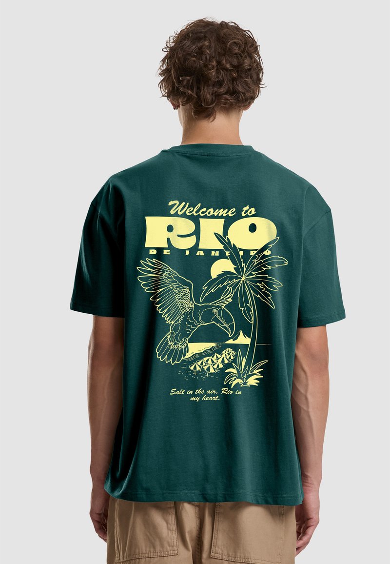 Personne aux cheveux bouclés portant un T-shirt vert avec le texte jaune « Bienvenue à Rio de Janeiro », toucan, palmier et parasols de plage dans le dos.