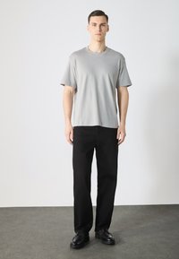 T-shirt grise à manches courtes en tissu doux, associée à un pantalon droit noir et à des chaussures noires, sur un fond neutre.