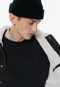 Beanie in maglia nera, felpa nera e giacca nera con interno in pelliccia bianca. Texture semplici, strati sovrapposti e bottoni metallici visibili.
