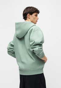 Νέος με κοντά σγουρά μαλλιά, ντυμένος με φαρδύ φούτερ σε χρώμα φασκόμηλου, τα χέρια στις τσέπες, γυρισμένος προφίλ σε απλό φόντο.