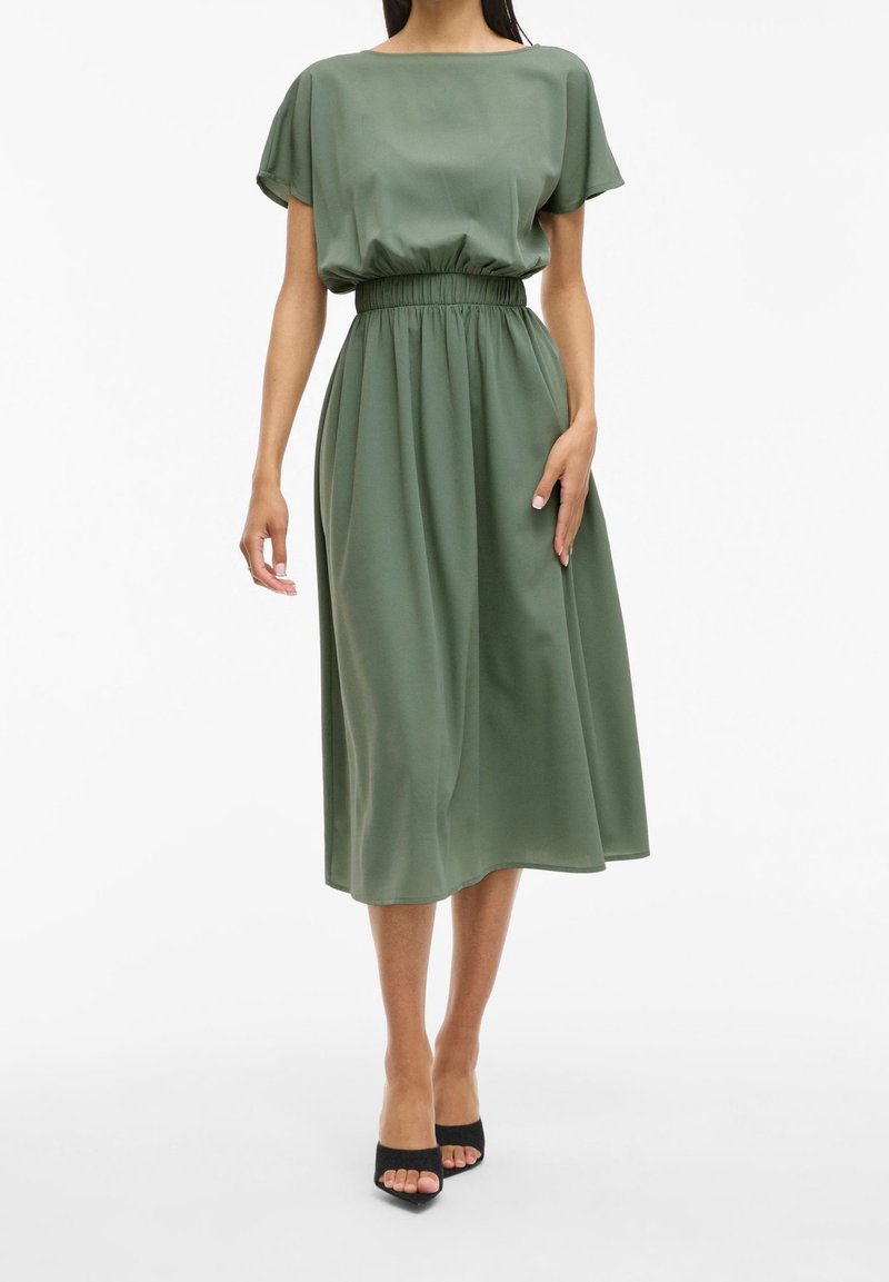 Femme portant une robe midi vert sauge à manches courtes et taille élastique, associée à des talons ouverts noirs, debout devant un fond blanc.