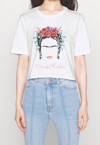 Bílý bavlněný tričko s grafickým potiskem tváře ženy zdobené květinami, s černými vlasy a textem "Frida Kahlo" v růžové barvě.