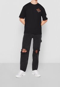 Camiseta de algodón negra con mangas cortas y un diseño gráfico naranja. Combinada con jeans negros desgastados y zapatillas blancas.