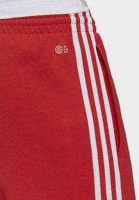 Pantaloni sportivi rossi in misto cotone con strisce bianche sui lati, fascia elastica in vita e un logo che riporta la scritta "FINE AI RIFIUTI DI PLASTICA".