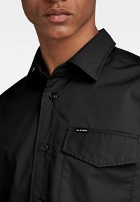 Camisa negra con un collar estructurado, cierre de botones y un único bolsillo en el pecho con el logo de G-Star. Textura suave, sin patrones.