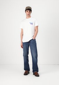 Hvid t-shirt med farverig grafik, blå denim jeans og brune sko. Modellen har også en brun ternet caps på. Enkel, afslappet outfit.