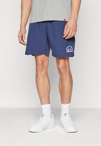 Navy blue shorts with a white Ellesse logo, featuring a smooth texture and angled side seams, paired with white sneakers and ribbed socks.