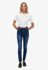 Camiseta blanca de algodón combinada con jeans de mezclilla azules de tiro alto. Cinturón trenzado marrón en la cintura, zapatos negros con borlas y un diseño recto.