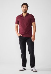 Bordeaux kortermet polo med glidelås i kragen, kombinert med svarte rettskårne jeans og hvite joggesko med svarte striper. Glatt stofftekstur.