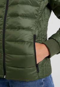 Gestepptes grünes Jacket mit horizontalen Nähten, schwarzem Reißverschluss und gerippten Bündchen; verfügt über eine seitliche Tasche mit Reißverschluss zur Aufbewahrung.