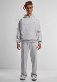 Pilkos spalvos treneriuko ir kelnių komplektas, pagamintas iš švelnios medžiagos, su gobtuvu, priekinėmis kišenėmis ir subtiliu logotipu siuvinėtu ant krūtinės ir šlaunies.