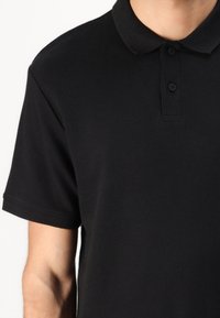 Zwarte polo met een ribbelstructuur, voorzien van een klassieke kraag en een knoopsluiting met twee zwarte knopen. Korte mouwen, minimalistisch ontwerp.