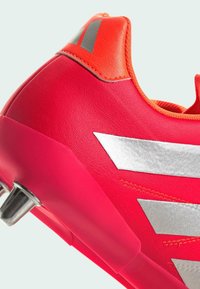 Bota de fútbol roja con exterior de cuero texturizado, acentos plateados y un llamativo talón naranja. Clavos de metal para tracción en la suela.