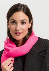 Fuksia roosa fliiskrae on mugavalt kaela ümber mähitud, pehme tekstuuriga, tumeda jope ja neutraalse tausta taustal.