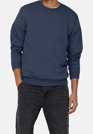 Mann, der ein schlichtes dunkelblaues Sweatshirt mit Rundhalsausschnitt und schwarze Jeans trägt, mit einer Hand in der Tasche vor einem einfarbigen Hintergrund.