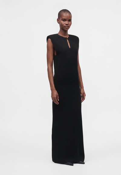Personne grande portant une robe maxi noire sans manches avec un décolleté en trou de serrure à l'avant et des chaussures noires pointues, debout devant un fond neutre.