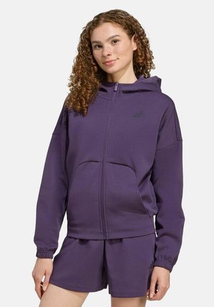 Giovane donna con capelli ricci che indossa una felpa con cappuccio viola con cerniera e pantaloncini abbinati, in piedi davanti a uno sfondo semplice.