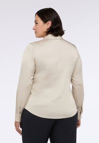 Blusa beige a maniche lunghe con texture liscia e leggera lucentezza, dotata di colletto e polsini con bottoni, abbinata a pantaloni scuri sartoriali.