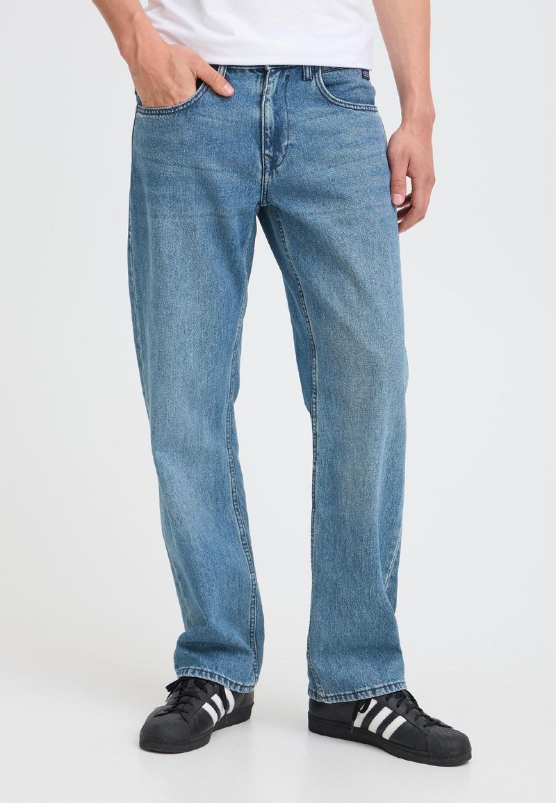 Mand iført lyseblå jeans med lige ben og sorte og hvide sneakers, stående med den ene hånd i lommen på en hvid baggrund.