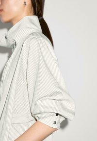 Lysegrå stribet jakke med høj krave, opslåede ærmer og knapdetaljer. Stoffet fremstår blødt med en subtil tekstur.