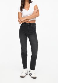 Jean noir taille haute à coupe slim, avec deux poches avant et une fermeture à bouton. Associé à un t-shirt blanc uni ajusté et des baskets.