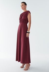 Dlhé bordové šaty s krátkymi rukávmi, zbieraným živôtikom a splývavou sukňou. Modelka má na sebe telové sandále na remienkoch a náramky na zápästí.