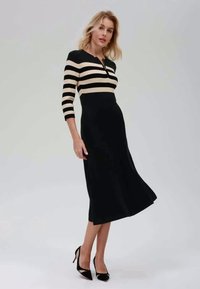 Čierne a béžové pruhované šaty s trojštvrťovými rukávmi, priliehavým korzetom a rozšírenou sukňou. Mäkka textúra a dĺžka po kolená. Čierne lodičky.