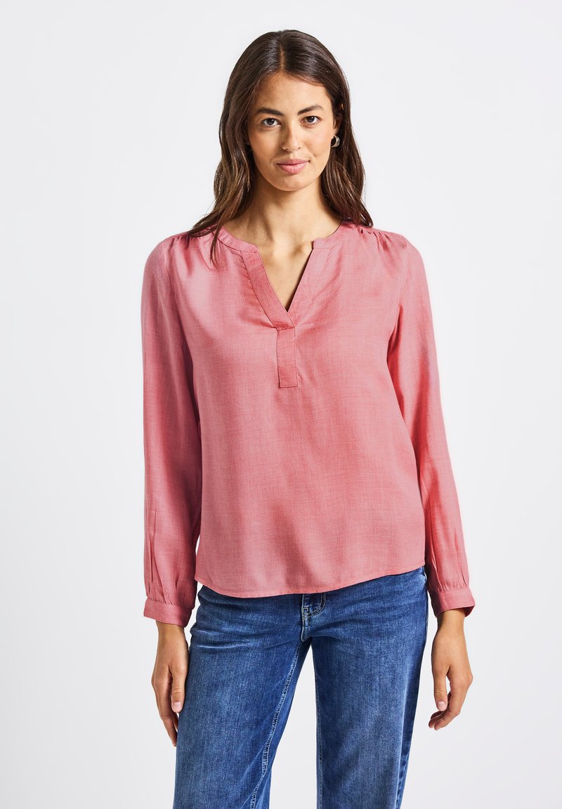 Blusa rosa chiaro a maniche lunghe con scollatura a V, delicate pieghe sulle spalle e vestibilità comoda, abbinata a jeans in denim blu.
