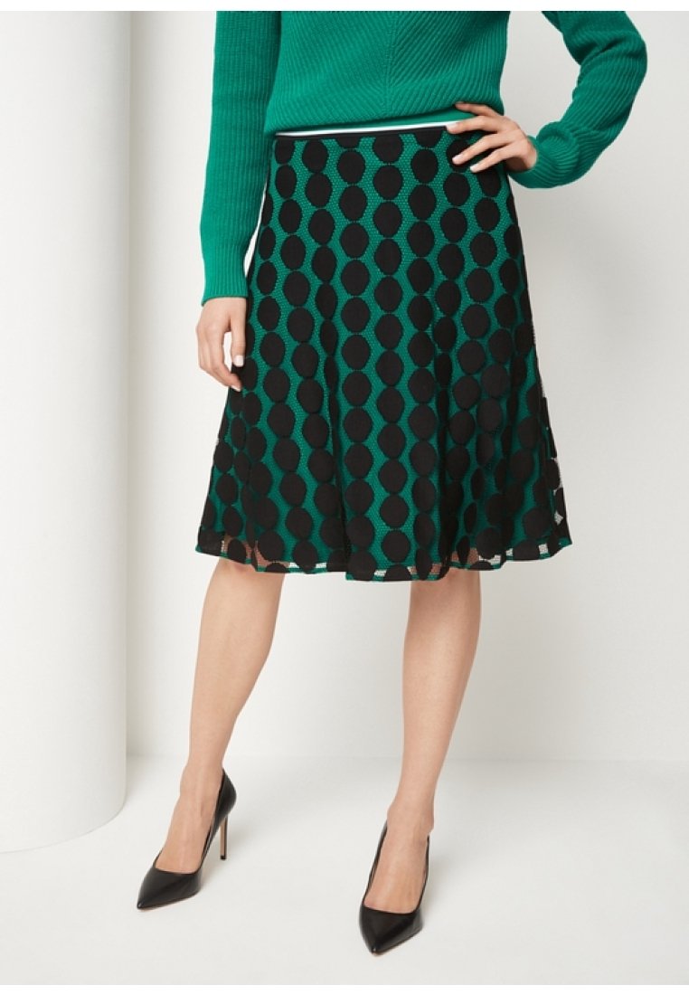 Mujer vistiendo un suéter verde, una falda A-line de lunares negros y verdes, y zapatos de tacón negros, de pie con la mano en la cadera.