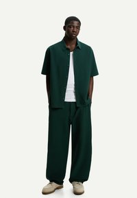 Dark green short-sleeve button-up shirt paired with wide-leg matching trousers, worn over a white tank top and beige shoes.