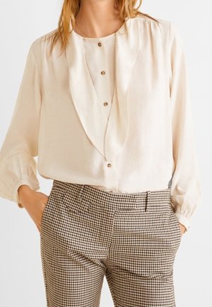 Femme portant un chemisier crème à boutons avec col drapé et manches longues, associé à un pantalon taille haute à motif écossais marron et blanc.