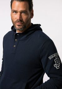 Sudadera con capucha de un color azul marino, con bolsillo frontal, cierre de tres botones y el bordado de "HERITAGE" en la manga en color gris. Textura de tela suave.