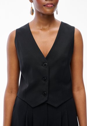 Kobieta ubrana w czarną bezrękawnikową kamizelkę z trzema guzikami i efektownymi kolczykami, pokazana od ramion do talii.