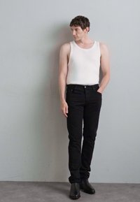 Μαύρα skinny τζιν με απαλό υλικό συνδυασμένα με λευκή αμάνικη μπλούζα. Το μοντέλο στέκεται μπροστά σε γκρίζο τοίχο, φορώντας μαύρες μπότες μέχρι τον αστράγαλο.