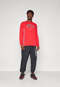 Man standing wearing red long-sleeve Bulls shirt, black Bulls pants, black and red sneakers, and red headband against plain white background.